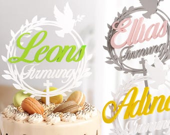 May include: Three white cake toppers with a dove and leafy wreath design. Each topper features a different name, "Leons", "Elias", and "Adina", with the word "Firmung" below each name.