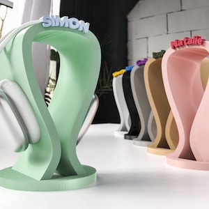 May include: A selection of colourful headphone stands, including green and pink, each with a name on top. A pair of silver headphones rests on the green stand. The stands are displayed on a white surface. The stands are in various colours.