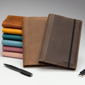 May include: Two brown leather journals with a black elastic band closure. The journal on the right has the name "JAMES" embossed on the cover. A stack of colorful leather journals is in the background.