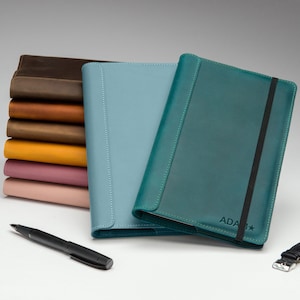 May include: Two leather-bound journals, one teal and one light blue, with a stack of six leather-bound journals in various colours behind them. A black pen and a wristwatch are also in the image.