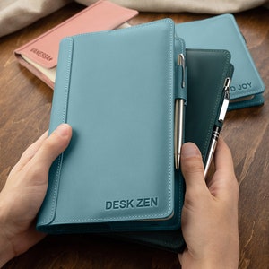 Könnte beinhalten: Ein tealfarbenes, ledergebundenes Notizbuch mit dem geprägten Text "DESK ZEN" auf dem vorderen Umschlag. Zwei silberne Stifte stecken in der Tasche des Notizbuchs.