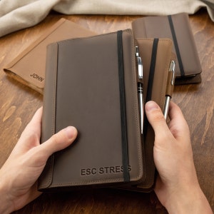 Può includere: Un diario in pelle marrone con chiusura a fascia elastica nera e un portapenne. Il diario ha il testo "ESC STRESS" in rilievo sulla copertina.