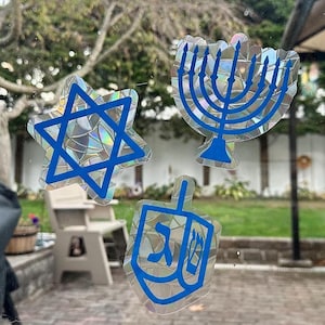 May include: Four blue and silver holographic stickers on a glass door. The stickers are of a Star of David, a menorah, a dreidel, and a Hebrew letter.