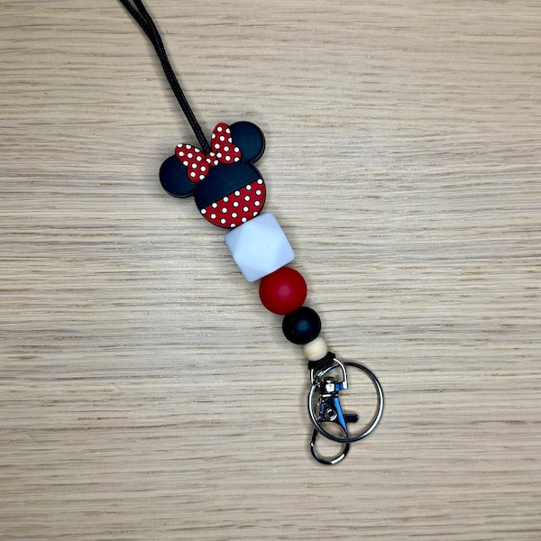 Könnte beinhalten: Ein Schlüsselband mit einem Minnie Mouse-Design. Die Oberseite hat eine schwarze und rote Schleife mit Tupfen und Ohren. Das Schlüsselband enthält weiße, rote, schwarze und Holzperlen sowie einen silberfarbenen Verschluss.