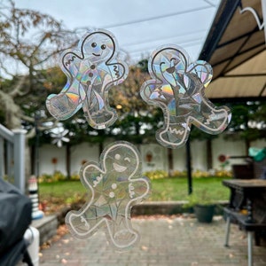 Puede incluir: Tres pegatinas de hombre de jengibre iridiscentes en una puerta de vidrio. Las pegatinas están hechas de un material transparente con un acabado holográfico. Los hombres de jengibre sonríen y tienen los brazos extendidos.