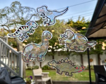 May include: Five iridescent holographic dinosaur stickers on a glass window. The dinosaurs are a T-Rex, a stegosaurus, a brachiosaurus, a triceratops, and a crocodile.