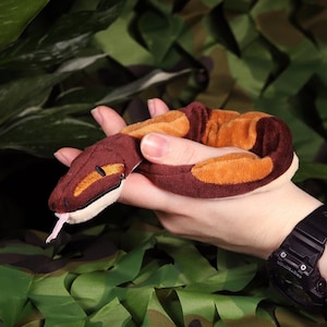 Op de afbeelding: Een bruin en oranje pluche slangen speelgoed met een roze tong. De slang wordt in de hand van een persoon gehouden.