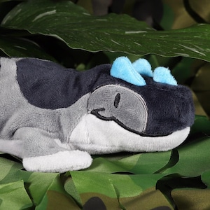 Puede incluir: Un dinosaurio de peluche gris y negro con púas azules en la cabeza. El dinosaurio está acostado de lado sobre un fondo de camuflaje verde.