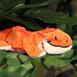 Peut inclure: Jouet en peluche orange et blanc en forme de lézard, couché sur un tissu camouflage vert et marron.