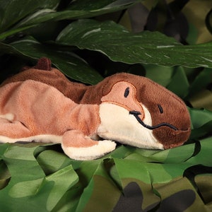 Puede incluir: Un dinosaurio de peluche marrón y crema acostado sobre una tela de camuflaje verde. El dinosaurio tiene una sonrisa y ojos cosidos.