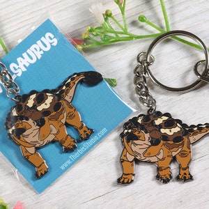 Puede incluir: Un llavero de metal con un dinosaurio Ankylosaurus de dibujos animados con detalles marrones, negros y blancos. El llavero está unido a un anillo plateado y una cadena plateada con un cierre de mosquetón. El llavero está empaquetado en una tarjeta azul con el texto "TRAV-SAURUS" y una dirección de sitio web.