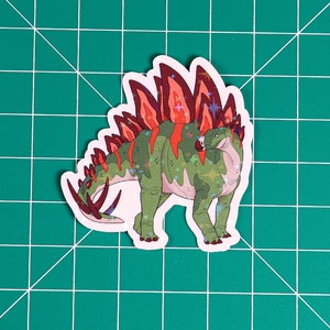May include: A green and red dinosaur sticker with a spiky back and glitter accents. The dinosaur is facing to the left and has a white outline.