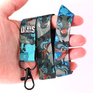 Può includere: Un laccio blu e verde con un moschettone in metallo nero. Il laccio presenta un motivo ripetuto di teste di dinosauro. Il testo "THE DEVILS DEMON" è stampato sul laccio.