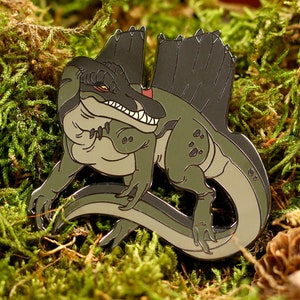 Può includere: Uno spillo in smalto verde e nero di un dinosauro dei cartoni animati con una testa grande e un collare. Il dinosauro è rannicchiato con la coda avvolta attorno al suo corpo.