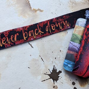 May include: A black and red wristband with the text "never back down" printed in gold. The wristband is paired with a card featuring a colorful dragon illustration.
