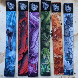 May include: Set of six colorful wristbands featuring different dinosaur designs. Each band has a black background with white text that reads "Teefers". The designs include a blue and white dinosaur, a red and white dinosaur, a purple and white dinosaur, a green and white dinosaur, an orange and white dinosaur, and a gray and white dinosaur.