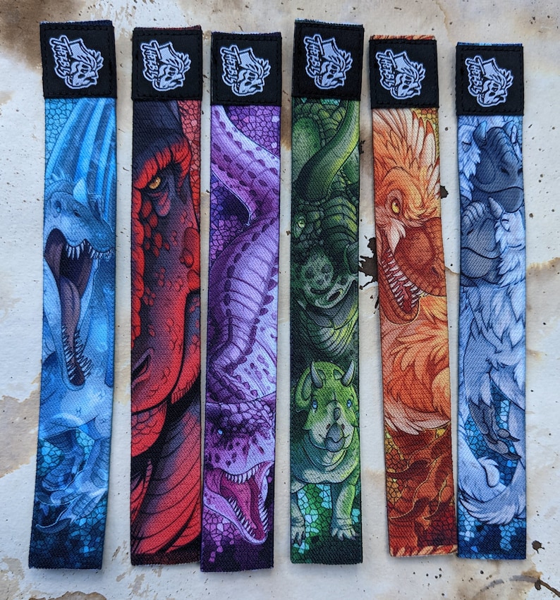 May include: Set of six colorful wristbands featuring different dinosaur designs. Each band has a black background with white text that reads "Teefers". The designs include a blue and white dinosaur, a red and white dinosaur, a purple and white dinosaur, a green and white dinosaur, an orange and white dinosaur, and a gray and white dinosaur.