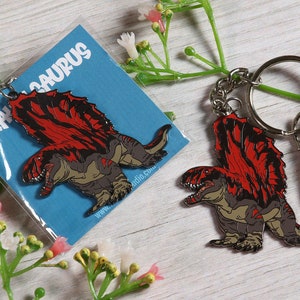 Może przedstawiać: Metalowy brelok w kolorze czerwonym i szarym przedstawiający dinozaura z kolczastym grzbietem. Brelok jest przymocowany do srebrnego kółka i srebrnego karabińczyka. Brelok leży na białym drewnianym tle.