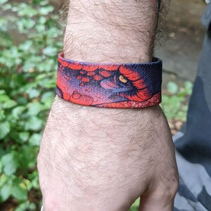 May include: A red and black wristband with a dragon eye design. The wristband is made of fabric and has a textured surface.