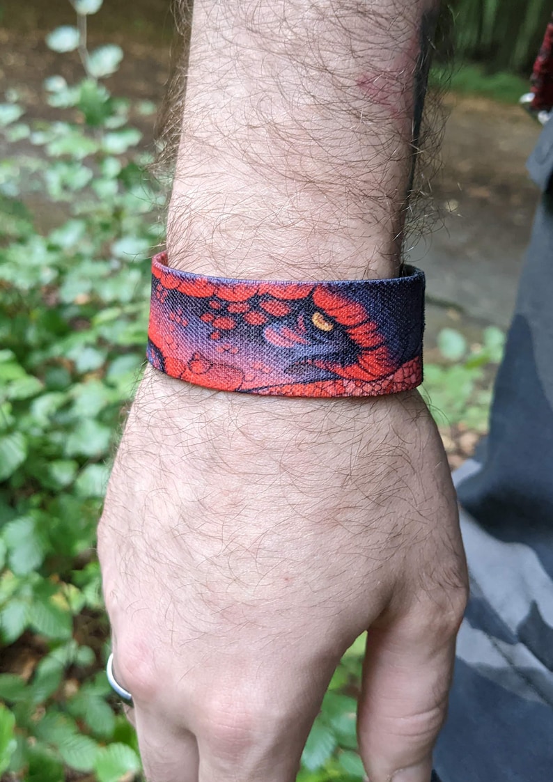 May include: A red and black wristband with a dragon eye design. The wristband is made of fabric and has a textured surface.