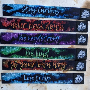 May include: Six colorful fabric wristbands with inspirational messages: Stay Curious, Never Back Down, Be Headstrong, Be Kind, Go Your Own Way, Love Truly. Each band has a black logo with the word "THELBIS" on it.