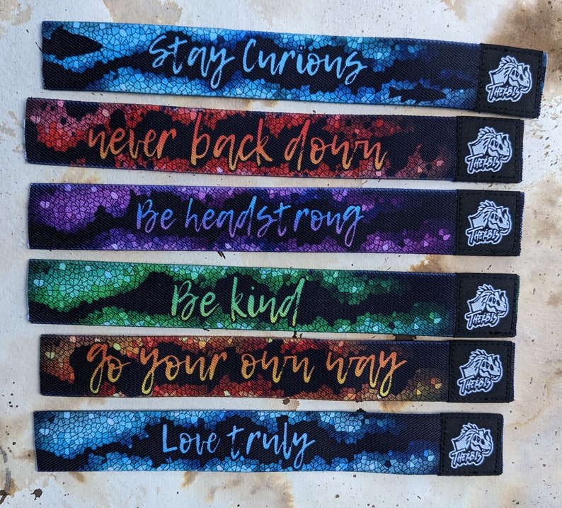 May include: Six colorful fabric wristbands with inspirational messages: Stay Curious, Never Back Down, Be Headstrong, Be Kind, Go Your Own Way, Love Truly. Each band has a black logo with the word "THELBIS" on it.