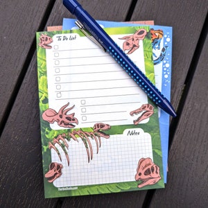 May include: A green and white notepad with a dinosaur theme. The notepad has a to-do list section with checkboxes and a notes section. The notepad has a dinosaur skeleton graphic and a blue pen.