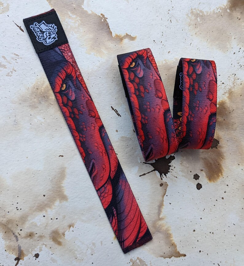 May include: Two red and black wristbands with a dragon design. The wristbands are made of woven fabric and have a black and white logo on one of the wristbands.