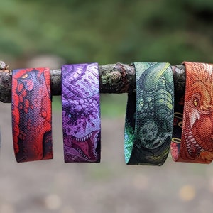 May include: Six colorful wristbands with different dragon designs. The wristbands are made of woven fabric and feature detailed illustrations of dragons with open mouths and sharp teeth. The colors of the wristbands are blue, red, purple, green, orange, and light blue.