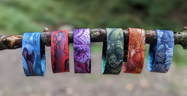 May include: Six colorful wristbands with different dragon designs. The wristbands are made of woven fabric and feature detailed illustrations of dragons with open mouths and sharp teeth. The colors of the wristbands are blue, red, purple, green, orange, and light blue.