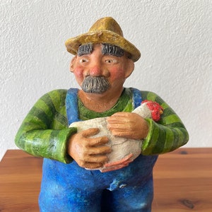 Keramikskulptur eines Bauern mit Overall, Hut und einem Huhn im Arm