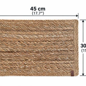 K&ouml;nnte beinhalten: Ein rechteckiges, geflochtenes Platzset aus naturfarbenen Fasern mit einem geflochtenen Rand. Das Platzset misst 45 cm in der Breite und 30 cm in der H&ouml;he. Eine kleine braune, rechteckige Lasche ist an der unteren rechten Ecke angebracht.