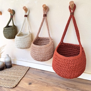 May include: Four hanging baskets in olive green, cream, dusty rose, and terracotta orange. Each basket is crafted from thick cord with a textured crochet stitch, featuring a single loop handle secured by a small brown leather tab at the top.