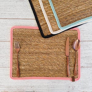 Puede incluir: Manteles individuales tejidos con ribetes de colores rosa, negro y azul claro. Un tenedor, un cuchillo y una cuchara de cobre están colocados sobre el mantel individual con ribete rosa. Los manteles individuales son de fibra natural.