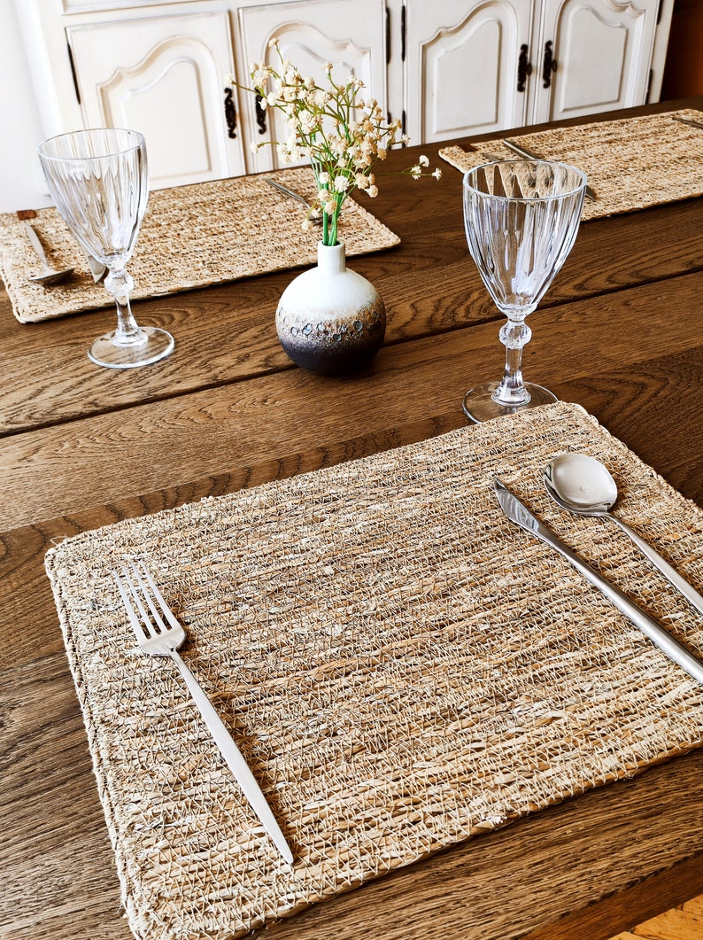 K&ouml;nnte beinhalten: Ein gedeckter Tisch mit rechteckigen, geflochtenen Stroh-Platzsets, Gabeln, Messern und L&ouml;ffeln aus Edelstahl, klaren, gerippten Weingl&auml;sern sowie einer kleinen, runden Keramikvase mit wei&szlig;em Oberteil und braunem Boden mit Trockenblumen.