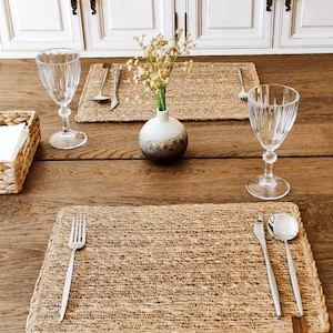 May include: A dining table set for two, featuring woven placemats, cutlery, and ribbed wine glasses. A small vase with dried flowers is placed between the settings. A basket with napkins is on the left.