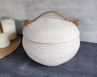 May include: A white round storage basket with a lid and a rope handle. The basket is made of woven rope and has a natural, rustic look.