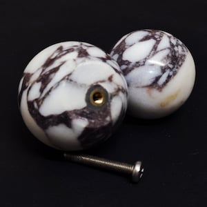 May include: Two marble cabinet knobs with a white and dark brown veined pattern. Each knob has a brass insert and is paired with a silver screw. The knobs are spherical and rest on a black surface.