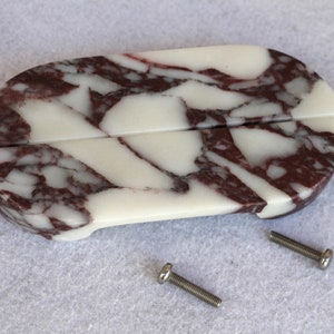 Peut inclure: Un plat ovale en marbre blanc et rouge avec deux fentes pour tenir les couverts. Le plat est composé de deux pièces qui sont assemblées. Deux vis argentées sont visibles sur la surface.