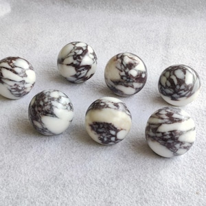 Puede incluir: Siete piedras esféricas lisas dispuestas en grupo. Cada piedra presenta una base de color blanco pulido con intrincados patrones de vetas de color púrpura oscuro a negro que crean una apariencia marmolada en toda la superficie de cada esfera.