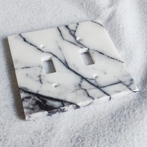 May include: A white marble light switch cover with black veining. The cover has two toggle switches.