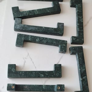 May include: A collection of six dark green marble cabinet pulls with a rectangular, U-shaped design. Each handle features a mottled, natural stone texture and includes two integrated brass-coloured metal mounting points on the underside.