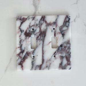 May include: A square, double light switch cover with a marble design. The cover has a white base with streaks of burgundy and grey. It features two rectangular cutouts for the switches and four screw holes.