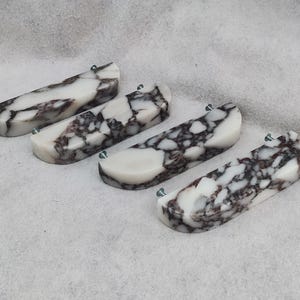 May include: Four rectangular cabinet pulls with a white and dark brown marbled pattern. Each pull has a curved top and is attached with small, light green screws. The pulls are arranged on a light gray surface.