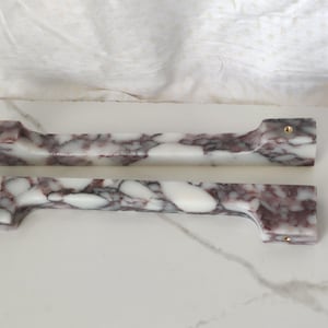 Könnte beinhalten: Zwei längliche Schrankgriffe aus weißem Stein mit ausgeprägter dunkelvioletter und kastanienbrauner Maserung. Jeder Griff hat ein rechteckiges Profil mit zwei kleinen, runden, goldfarbenen Metallschraubeneinsätzen an den Enden zur Montage.