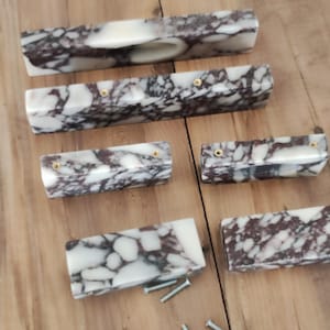 Peut inclure: Un ensemble de six poignées de placard rectangulaires présentant un motif en marbre blanc et violet foncé poli. Chaque poignée possède une finition lisse et brillante avec des inserts de vis en métal doré visibles pour la fixation.