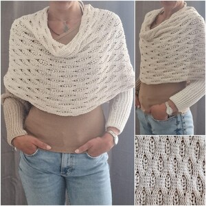 May include: Cream-colored knitted shawl with a cowl neck and openwork design. The shawl is worn over a tan top and light blue jeans. The knitted pattern is visible in a close-up shot.