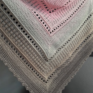 Op de afbeelding: Een handgemaakte gehaakte sjaal in roze, grijze en beige tinten. De sjaal heeft een gedetailleerd openwerk ontwerp met een geschulpte rand. De kleurovergang verloopt van roze naar grijs naar beige.