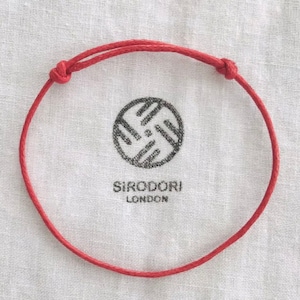 May include: Red adjustable bracelet with knotted ends. The bracelet is displayed on a white cloth with a black circular logo and the text "SIRODORI LONDON".