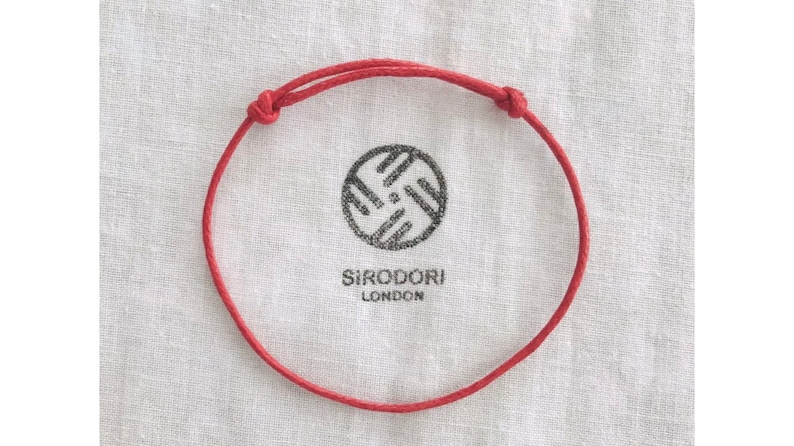 May include: Red adjustable bracelet with knotted ends. The bracelet is displayed on a white cloth with a black circular logo and the text "SIRODORI LONDON".